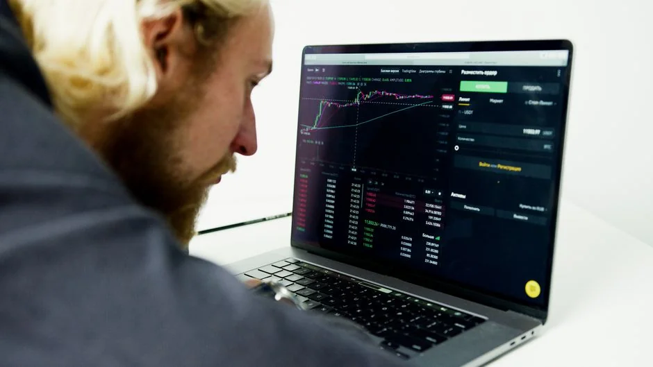 Hands using smartphone beside laptop with stock charts, showcasing digital trading.